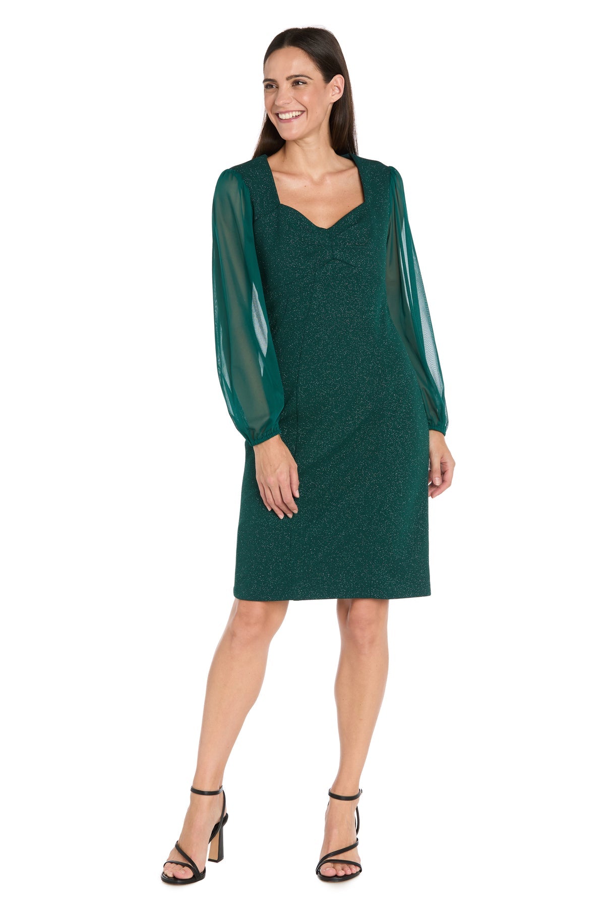 A woman stands smiling in the R&M Richards 4331P petite short green party dress with sheer long sleeves, styled with black high-heeled sandals.